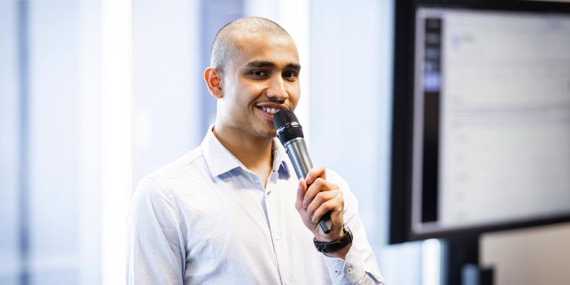 A young man speaks to a group out of shot using a microphone