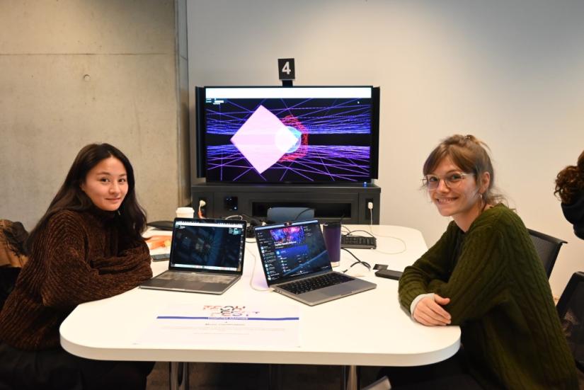 Two girls sit in front of laptops and display showing graphics