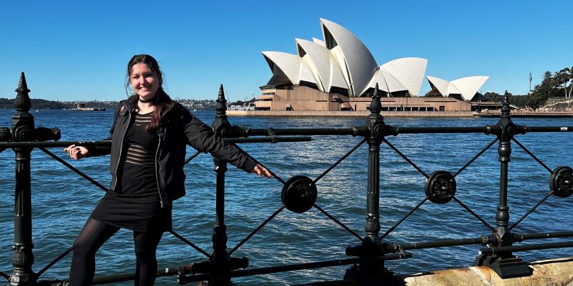 Agata stands left of the Opera House with her arms spread out, leaning against black railing. Agata is wearing all black and it’s a clear, sunny day.