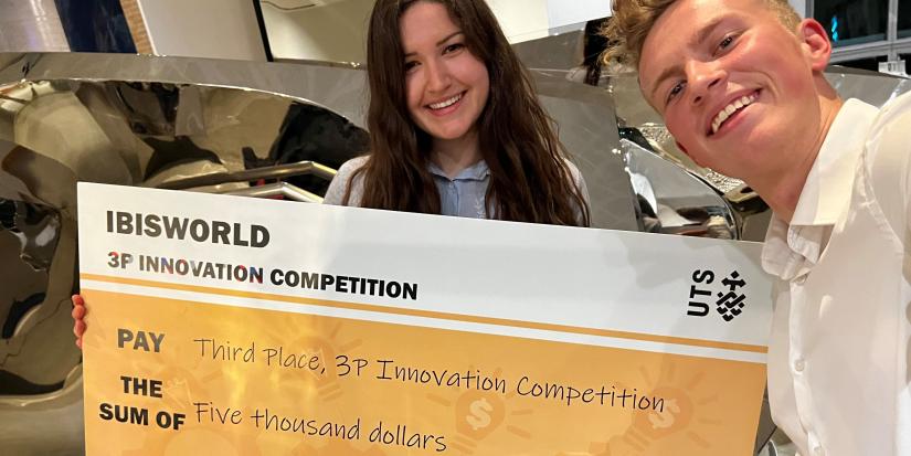 Thomas (right) takes a selfie with Agata, who is holding a large novelty check that reads "IBISWORLD 3P Innovation Competition". Agata is brunette with wavy hair and Thomas is blonde with a short cropped hairstyle.