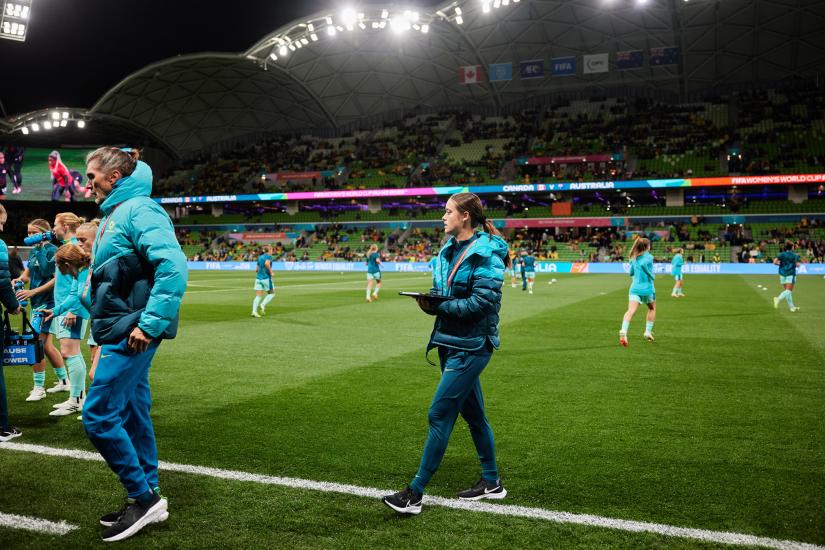 A woman dressed in a teal uniform holds a clipboard on the sidelines of a football pitch, players training in the background.