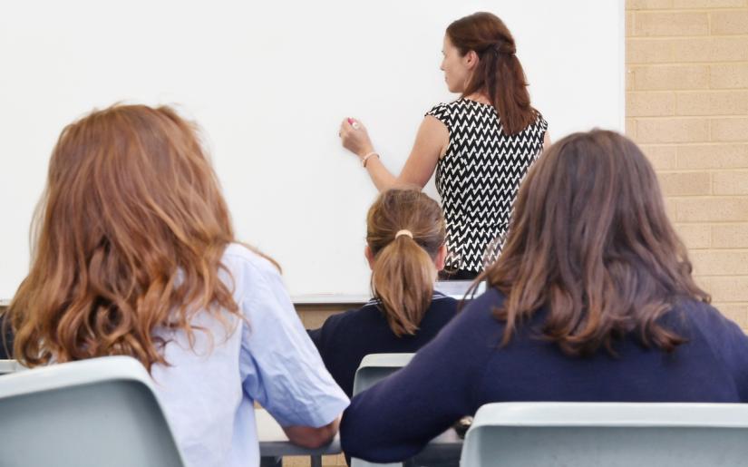 Three young students sitting in class look towards a teacher writing on a whiteboard.