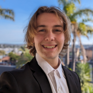 A headshot of Callum Burke from the chest up. Callum has brown hair and is wearing a black blazer with white collared shirt. The background is an out of focus bright blue sky with palm trees.