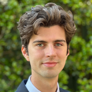 A headshot of Cooper Crellin from the shoulders up. Cooper has dark brown hair and is wearing a navy blazer with a stripey blue tie. The background is an out of focus green hedge.