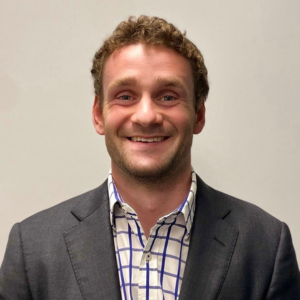 A headshot of Matthew Dutaillis from the chest up. Matthew has cropped brown hair and is wearing a dark grey blazer with a white and blue patterned shirt. The background is plain grey.