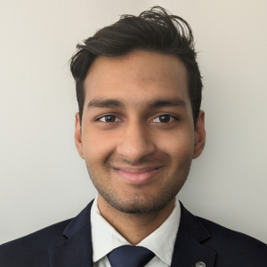 A headshot of Sai Muthukumar from the shoulders up. Sai has cropped dark brown hair and is wearing a navy blazer with a navy tie. The background is plain grey.