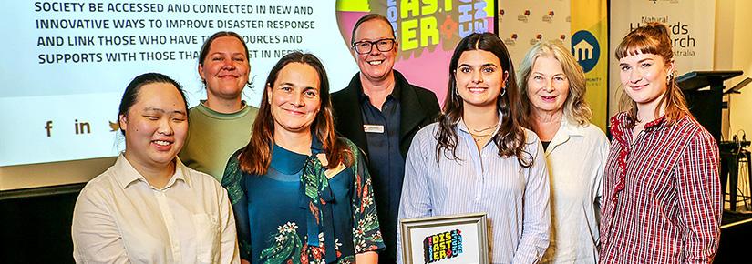 6 people of mixed genders and ages stand in front of a screen. A female UTS students holds the award won by her team for the Disaster Challenge.