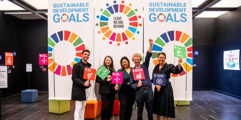 Five members of the UTS BUILD and Sustainability programs stand smiling in front of three vertical banners that read “SUSTAINABLE DEVELOPMENT GOALS”. The banner features multicoloured circular graphics. The members hold orange, green, pink and blue signs that correlate with the United Nations 17 Sustainable Development Goals.