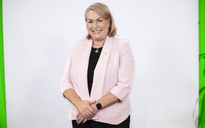 Dr Michele Bruniges AM stands against a white wall wearing a pink jacket and a black shirt, smiles at camera.