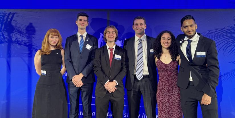 Six students - four male, two female - stand against a rich blue backdrop. They are wearing black suits and dresses, smiling. From left to right: Madeleine McWlliam; Cooper Crellin; Callum Burke; Matthew Dutaillis; Samantha Morris; and Sai Muthukumar.