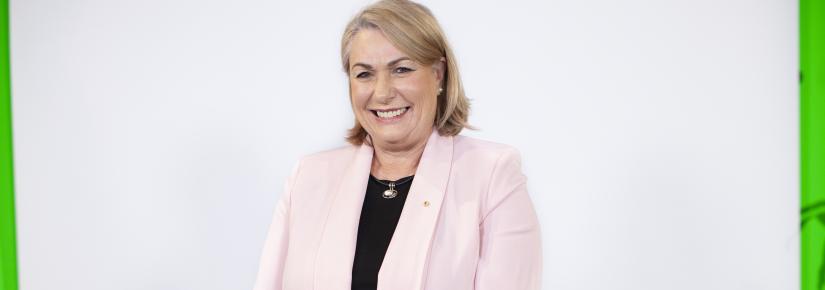 Dr Michele Bruniges AM stands against a white wall wearing a pink jacket and a black shirt, smiles at camera.