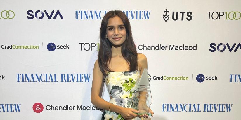 Arya Lohar standing in front of sponsors logos with AFR Award in hand.