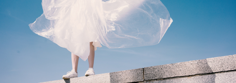 Feet in sneakers standing on a wall in front of blue sky, person appears to be wearing a large piece of plastic that is floating in the breeze.