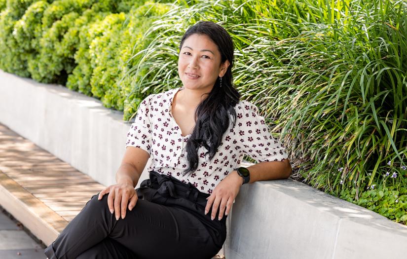 Woman seated outside under trees. She is resting her left arm on a a wall and she is smiling and looking at the camera. Soph-ea is wearing a white top with a black pattern and black pants. She has her hair tied in a low ponytail on the side