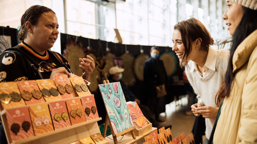 Two women interact with a stallholder at an art fair. 