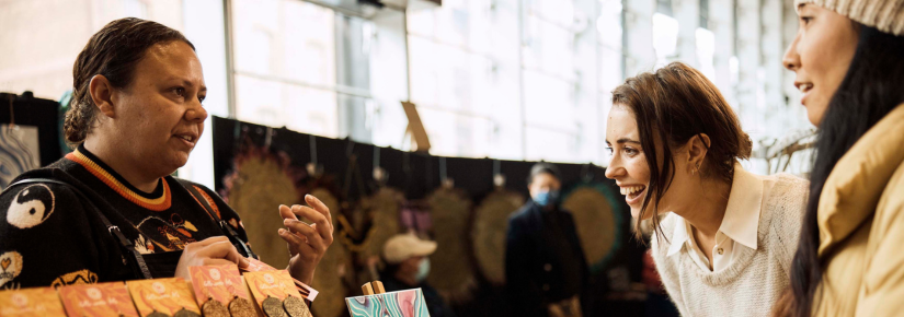 Two women interact with a stallholder at an art fair. 