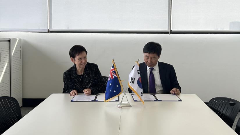An Australian female and a South Korean male each sign a document behind their respective national flags.