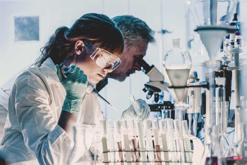 Female research in the foreground wearing safety goggles and a research lab coat. She is holding a syringe and putting a liquid into a test tube. Male researcher in background looking into a microscope.