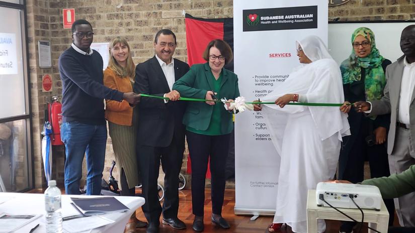 Sudanese cultural day celebration: Group of people holding ribbon, being cut to launch SAHWA