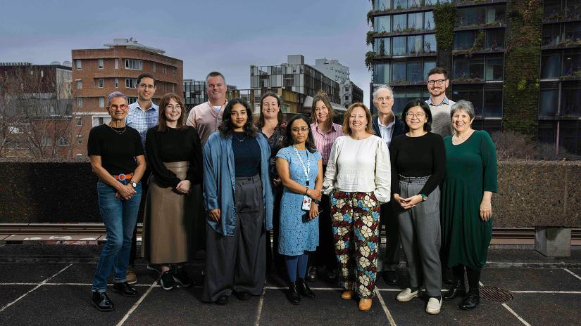 Group photo of Accessibility Service staff – 13 staff standing and smiling. Taken on level 6 of the UTS Tower with a background of various buildings from Broadway including the Central Park building with vertical gardens cascading down the side.