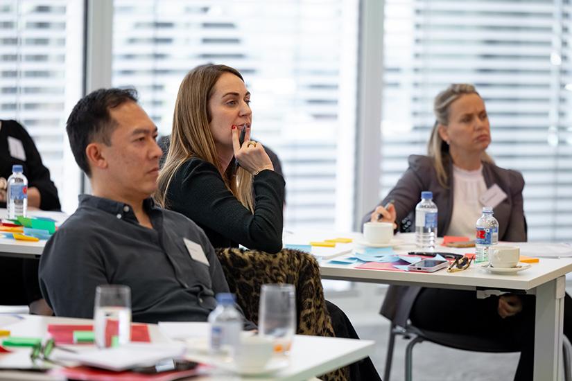 Photo of three alumni concentrating on something at the front of the room. The woman in focus has her hand up to her face with a pen in it.