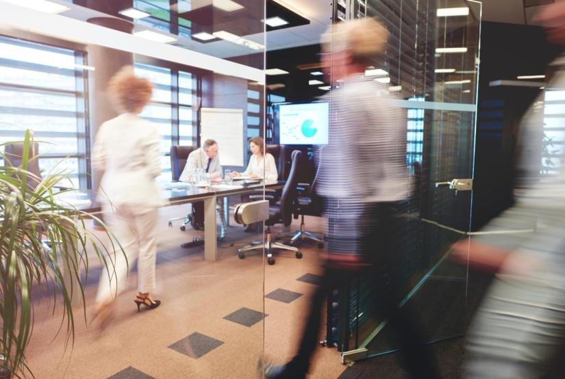 Stock picture of an office with a man and woman at a desk and two blurred moving figures of women