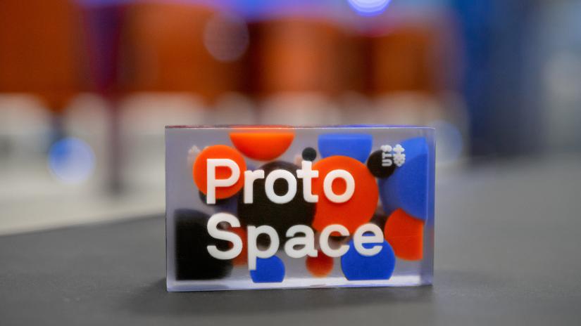 Coloured 3D printed part in focus, featuring ProtoSpace logo in white, many coloured spheres around, set in a clear block, with many printers out of focus in background.