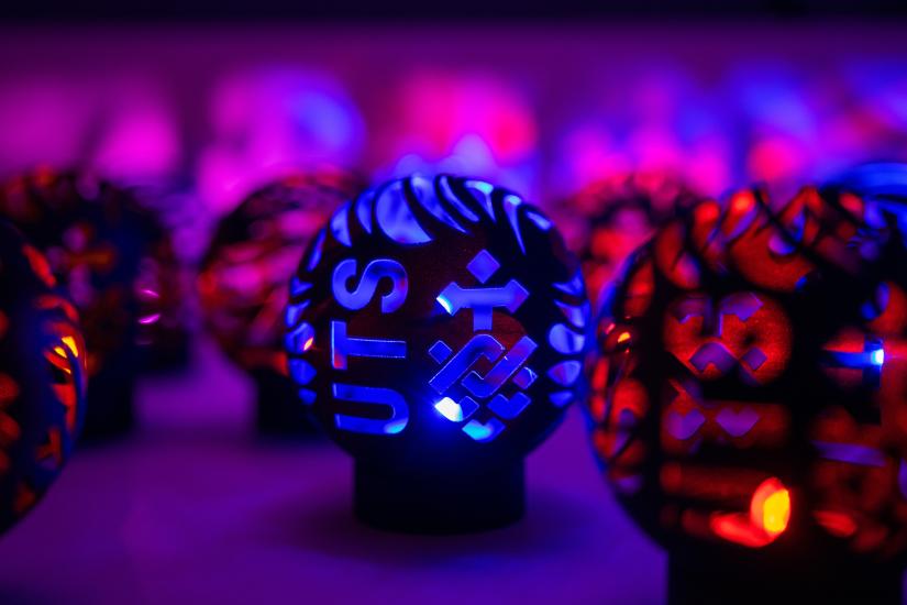 A spherical, black, hollow 3D printed part with blue LED lighting from within, featuring the UTS logo, among other unfocused parts in background.
