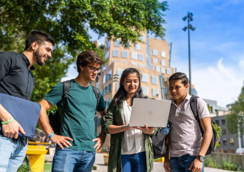 Four students looking at a laptop outside with the Business building in the background