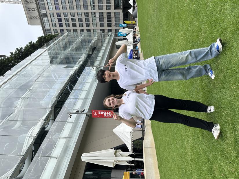 Two students are standing on the grass at Alumni Green in front of Building 2. They are both wearing white UTS t-shirts