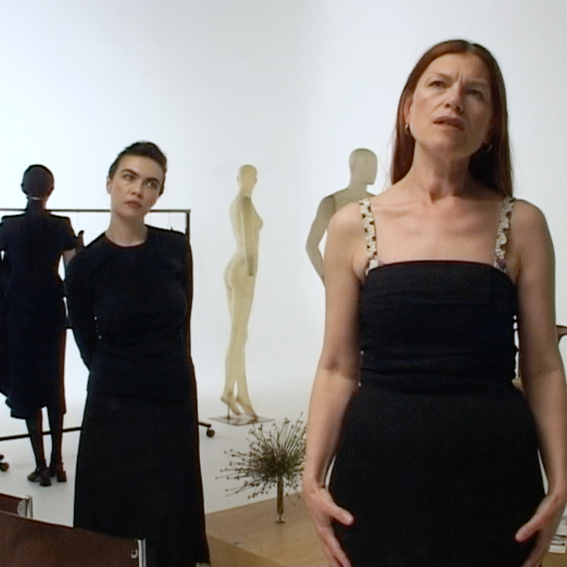 Two women in black dresses stand in a minimalist gallery space with mannequins and clothing racks behind them, creating a fashion-focused, contemplative scene.