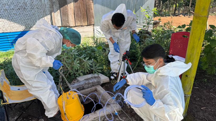 Two researchers dressed in hazmat suits and masks pull up a bucket of water from a hole in the ground.