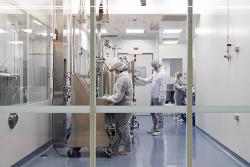 Lab technicians in protective clothing, masks and gloves work on high-tech equipment behind a glass viewing window