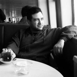 Seref Isler sitting at a cafe table drinking Turkish coffee. Image in black and white.