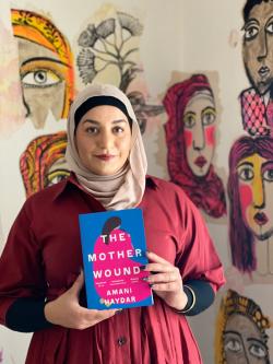 image of woman standing in front of artworks of women's faces. The woman is wearing a dark red shirt, headscarf and is holding a copy of a book. the book cover is blue and pink with an illustration of a woman and the text reads 'The Mother Wound' and 'Amani Haydar'.