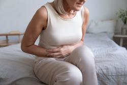 Older woman with blonde hair and cream sleeveless top sits on edge of bed clutching her tummy