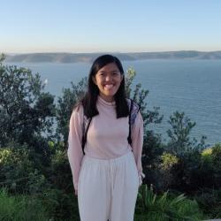 Freyja stands smiling in front of on a bushwalk overlooking the ocean.