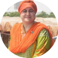 Aisha sitting outside in traditional dress, wearing a orange hat and a green and orange sari. She is smiling and looking at the camera.