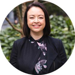 Belinda Bentley standing outside in front of a garden. Belinda has shoulder length black hair and is wearing a black jacket with a black and purple top underneath. She is smiling and looking at the camera.