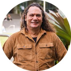 Clarence standing in front of a garden on the UTS Alumni Green. Clarence has shoulder length curly hair and is wearing a brown button up shirt. He is smiling and looking at the camera.