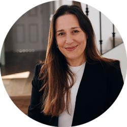 Nicky Sparshott smiling and looking at the camera. Nicky has long brown hair and she is wearing a black suit jacket with a white tshirt.