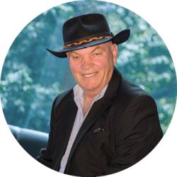 Professor Jack Beetson sitting in front of a window wearing a black hat with a purple business shirt and a black suit jacket. He is smiling and looking at the camera.