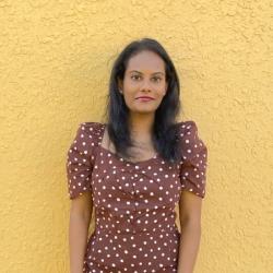 Woman wearing white polka dot dress on yellow background looks into camera.