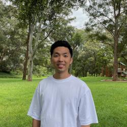 Man in white t-shirt stands in front of trees and grass.