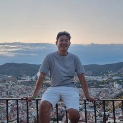 A man sits on a railing with a city scape behind him. 