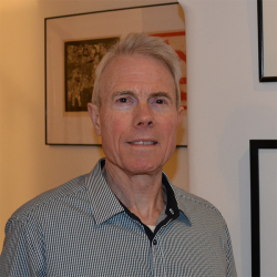 Stephen Peterson has short grey hair and he is wearing a button up shirt. He is standing in front of a gallery wall of paintings.