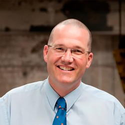Professor David Currow is wearing a pale blue buttoned shirt with a bright blue tie. He is wearing glasses and is smiling at the camera.