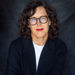 A picture of Danielle Woolley - Dark hair, round glasses, red lipstick wearing a black suit jacket with a white shirt.