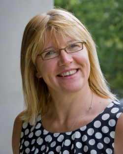 Photo of Sara Wilkinson smiling and wearing a black and white polka dot top with a grey wall and trees behind her.