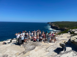 A group of international students stand on the coast with the blue sky and ocean in the background